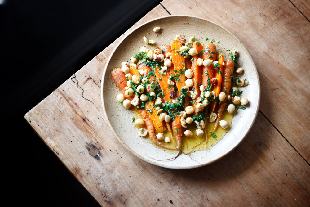 Whole roasted carrots topped with chopped herbs and toasted hazelnuts, served on a ceramic plate with a drizzle of olive oil.