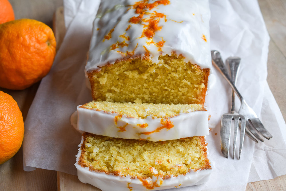 Glazed Seville orange loaf cake topped with white icing and grated orange zest. Sliced and served on chopping board with dessert forks and fresh oranges.