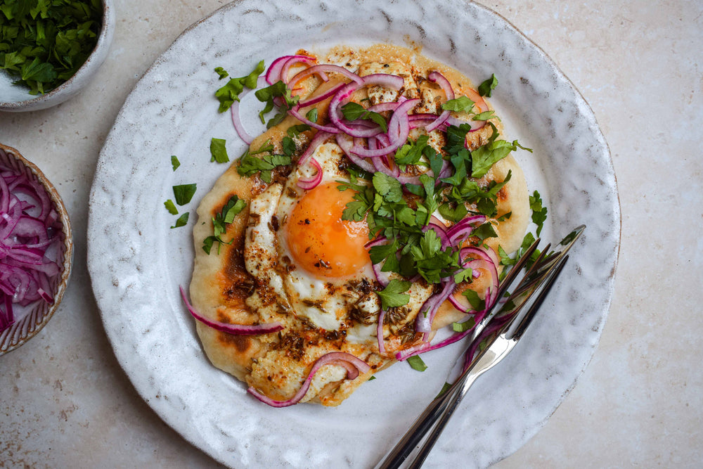 Fried egg in spiced butter topped with green garnish and red onion, served on a warm, rustic flatbread on a white plate